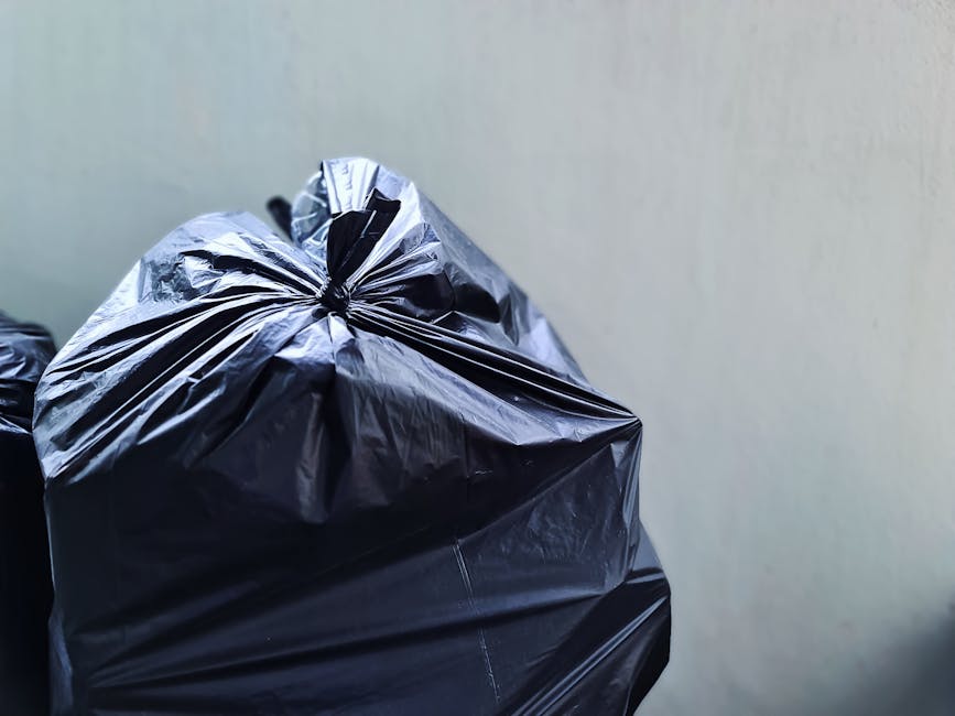 A large outdoor waste collection area shows an overflowing metal recycling bin filled with mixed paper and cardboard, with various tangled materials spilling onto the surrounding pavement. To the righ
