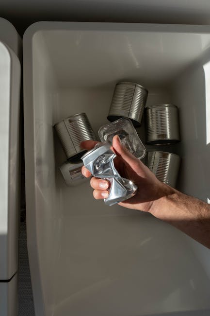 A hand holds a crumpled clear plastic water bottle above a beige metal recycling bin labeled 'METAL,' set against a dark background with red fabric curtains. The bottle appears partially crushed, with