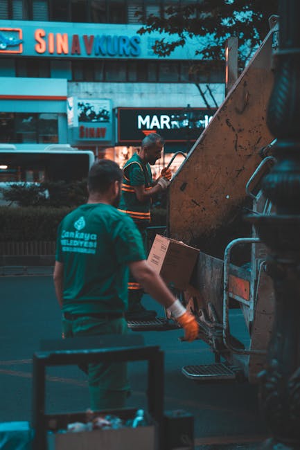 Two waste collection workers stand next to a large, rusted metal skip on a city street during dusk, with illuminated shop signs visible in the background. One worker, wearing a teal uniform with the t