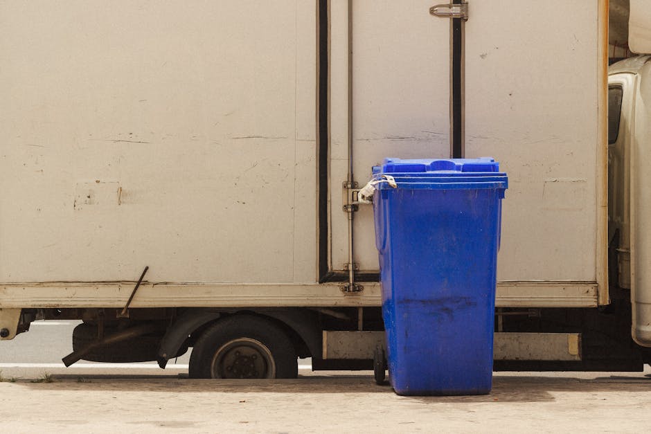 Two waste collection workers stand next to a large, rusted metal skip on a city street during dusk, with illuminated shop signs visible in the background. One worker, wearing a teal uniform with the t