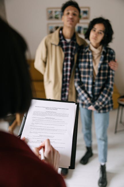 A woman holding a clipboard with a document facing two young adults, a man and a woman, who are standing together indoors in front of a blurred background with framed pictures. The woman is partially
