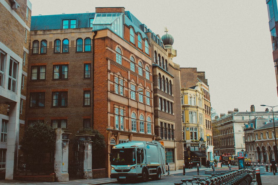 A city street scene featuring a row of multi-story historic buildings with brick and ornate window details, situated on the left and right sides of the image. In the foreground, a light grey commercia