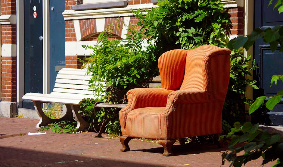 Three vintage armchairs positioned in a row on a paved outdoor surface in front of a weathered wooden wall with a corrugated metal roof above. The left armchair features a wooden frame with upholstere